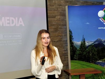 Woman presenting on social media in a conference room, holding a clicker, with a golf course image in the background and a projection screen beside her.
