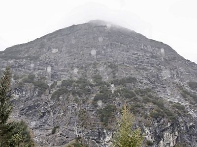 A large rocky mountain with a misty peak, surrounded by scattered greenery and cloudy sky, creating a dramatic natural scene.