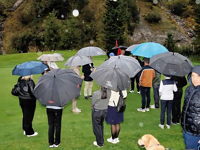 A group of people holding umbrellas stands on a grass field in the rain, surrounded by trees. A person looks on, and a dog is visible in the foreground.