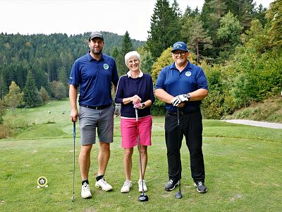 Three people on a golf course: two men in blue shirts and a woman in pink shorts, all with golf clubs, standing on green grass with a forest backdrop.