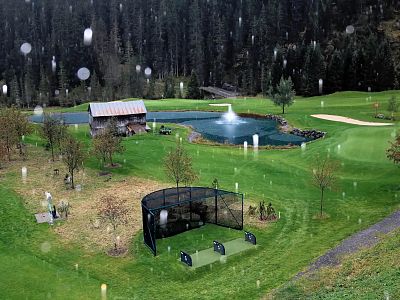 A rainy golf course with a cabin, pond, and forest backdrop. Green grass, scattered golf nets, and gentle raindrops create a peaceful, natural scene.