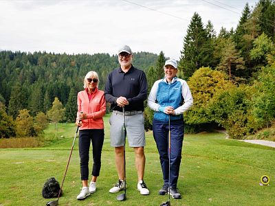 Three golfers stand on a grassy course with trees in the background, holding clubs and wearing golf attire on an overcast day.