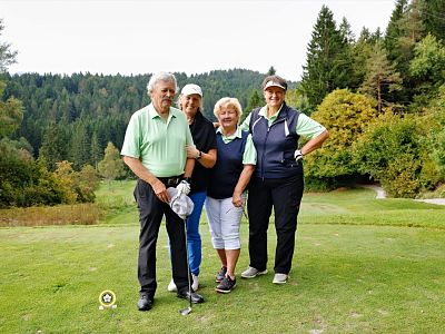 Four people stand on a golf course with trees in the background, wearing casual golf attire. One holds a club, and they seem to be enjoying a sunny day.
