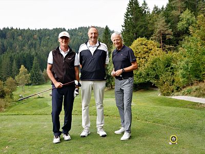 Three men stand on a golf course, holding clubs, with a backdrop of lush green trees and a clear sky, wearing golf attire and smiling at the camera.