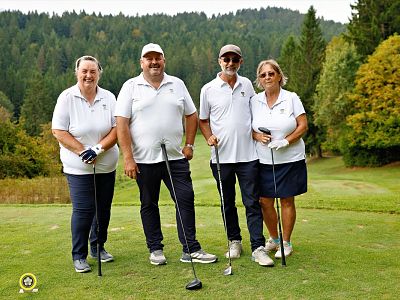 Four individuals in white shirts, two men and two women, stand on a golf course holding golf clubs. Trees and green foliage are visible in the background.