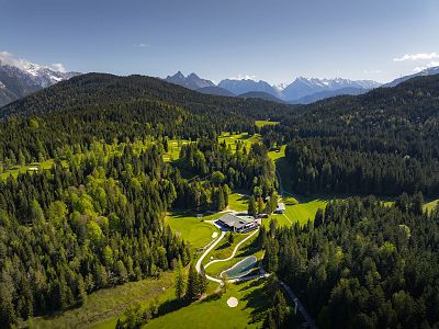 Aerial view of green mountains, dense forests, and a modern house with a winding road. Clear blue sky above the lush landscape.