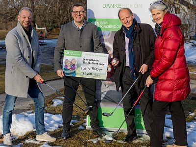 Group of four people outdoors with a large check and golf clubs, on a snowy lawn, indicating a charitable event.
