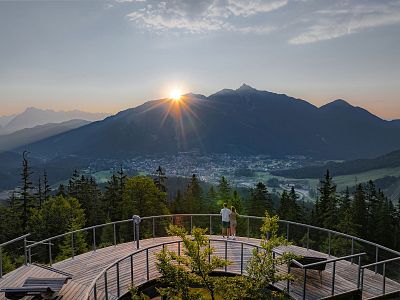 Sunrise over mountains seen from a wooden viewing platform with trees and a town visible. The sky is clear with some clouds, creating a peaceful ambiance.