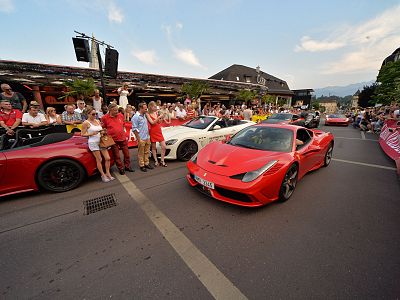 A street scene with several red and white sports cars and a crowd of people watching, set in a lively environment with buildings and a clear sky in the background.
