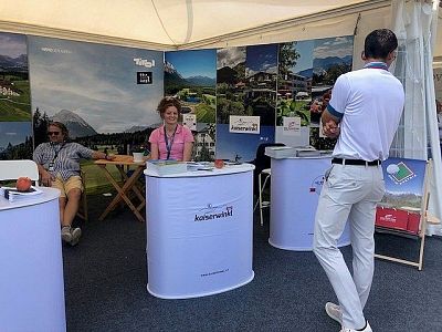 A man in white talks to seated staff at a tourism booth with scenic posters and brochures, set up under a canopy at an outdoor event.