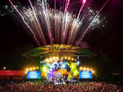 Fireworks explode over a colorful outdoor concert stage at night, with bright lights, a cheering crowd, and musicians performing under a tent-like canopy.