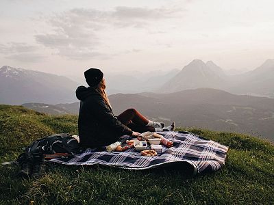 A person sits on a picnic blanket overlooking a scenic mountain view, surrounded by greenery and a cloudy sky.