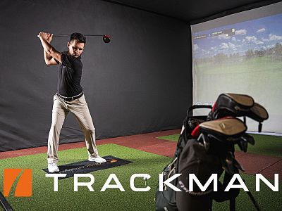 Man practicing a golf swing indoors with a simulator, featuring a golf mat, projection screen showing a course, and golf bag nearby.