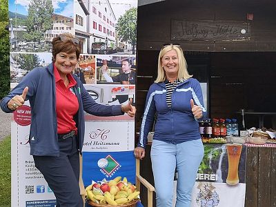 Zwei lächelnde Frauen stehen vor einem Holzkiosk mit Getränken und Plakaten. Sie zeigen den Daumen hoch und tragen Freizeitkleidung.