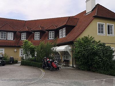 A charming country house with red roof tiles, yellow walls, and a neatly kept garden. Several golf bags and a buggy are visible near the entrance.