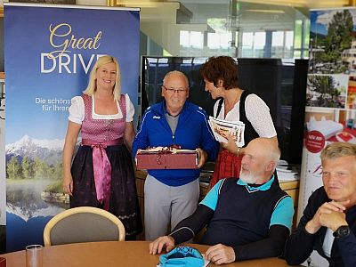 A woman in a dirndl presents a gift to a man, with others seated nearby, during a gathering in a room with a "Great Drive" banner.