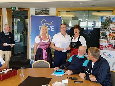 Eine Gruppe von Menschen steht in einem Raum, einige in traditioneller bayerischer Kleidung. Im Hintergrund sind Poster und eine Glastür zu sehen.