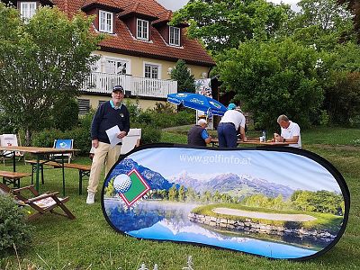 Golf event with participants near a house; promotional banner shows a scenic course.