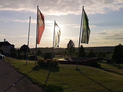 Three flags wave against a sunset backdrop in a serene park. The sun creates a warm glow on the landscape with trees and open fields in view.