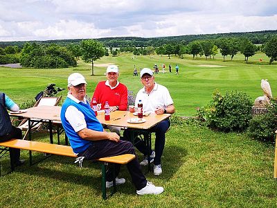 Three golfers sitting at a table with drinks on a green golf course, with trees and fairway in the background.
