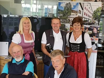 A group of five people in Bavarian attire, with women in dirndls and men in jackets, posing indoors with a poster of a building in the background.