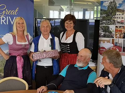 People in traditional Bavarian attire gather indoors. One holds a decorative box. It's a casual, celebratory setting with posters in the background.