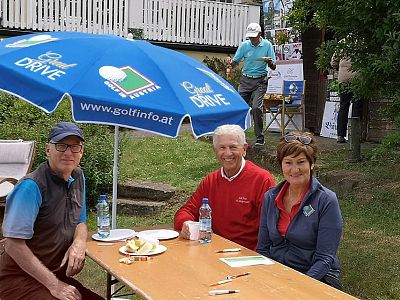 Three people sit at an outdoor table under a branded umbrella, with grass and trees in the background, suggesting a golf event setting.