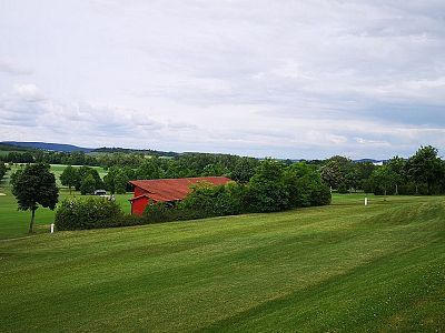 A green field with a red barn, surrounded by trees under a cloudy sky, creating a tranquil rural scene.