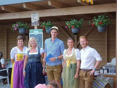 Five people in traditional Bavarian clothing, smiling outdoors with wooden backdrop and flowers. Includes women in dirndls and men in lederhosen.