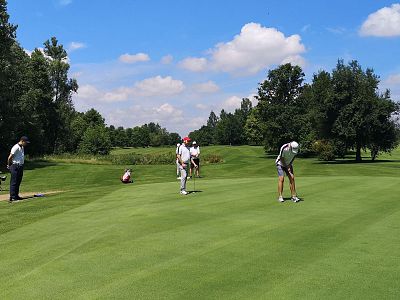 Several golfers on a lush green golf course, one putting the ball while others watch, surrounded by trees under a blue sky with fluffy clouds.