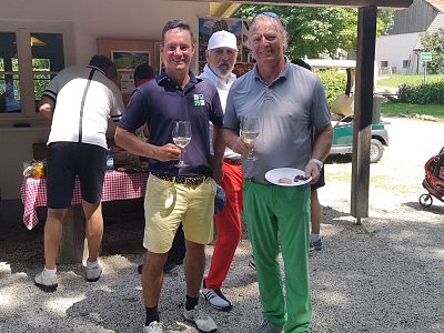 Two men in golf attire holding wine glasses, smiling outdoors at an event. Others in the background by a table with drinks and snacks, near a golf cart.