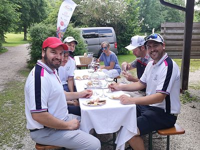 Six people sitting at a white tablecloth-covered table outdoors, enjoying a meal surrounded by greenery. They are in casual attire, smiling and chatting.