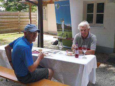 Two men sit at an outdoor table set with a white cloth, enjoying a meal. A wooden fence and a house with windows are in the background.