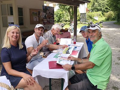 Eine Gruppe von fünf Personen sitzt an einem Tisch im Freien, umgeben von grüner Natur. Sie scheinen Dokumente oder Broschüren zu betrachten und lachen.