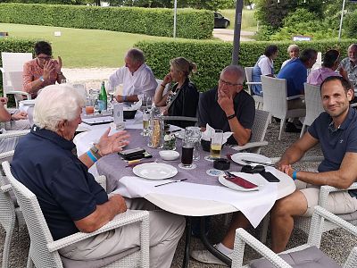 A group of people sitting at a table outdoors with plates and drinks, surrounded by greenery. Casual atmosphere, with a focus on socializing.