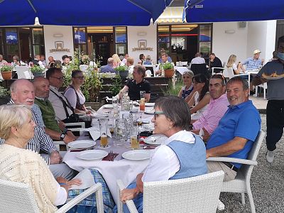A lively outdoor restaurant setting with a group dining at a long table. People are conversing and enjoying their meals under blue umbrellas.