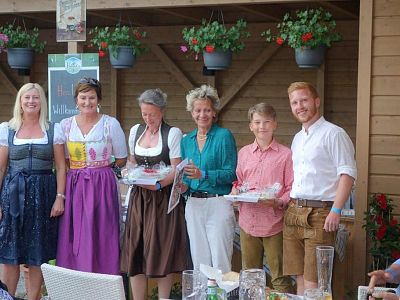 Six people in traditional Bavarian attire stand smiling, some holding items, in front of a wooden backdrop with potted flowers hanging above.