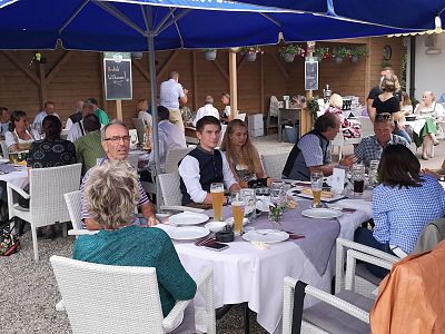 Gruppe von Menschen sitzt unter blauen Sonnenschirmen in einem Außenbereich eines Restaurants. Die Tische sind gedeckt, es herrscht eine gesellige Atmosphäre.