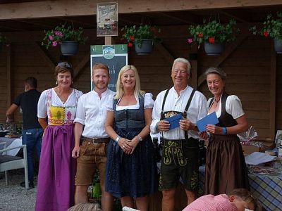 A group of people in traditional Bavarian clothing stand smiling at an outdoor event with wooden and floral decorations visible in the background.