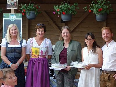 Eine Gruppe von fünf Personen steht lächelnd vor einer Holzwand. Einige tragen traditionelle bayerische Trachten. Blumenkästen hängen über ihnen.