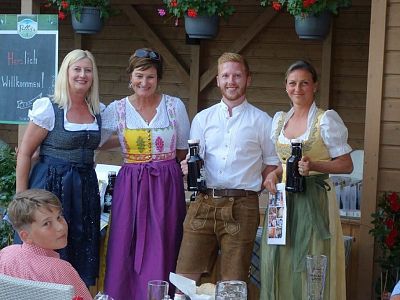 Four adults in traditional Bavarian clothing posing with drinks, while a boy in the foreground gazes at the camera. Wooden decor and plants in the setting.