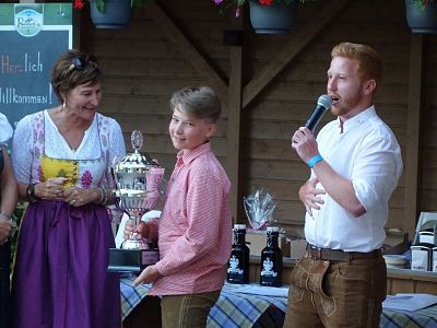 Boy receiving a trophy from a woman while a man speaks into a microphone at an event with rustic decor. The atmosphere is casual and festive.