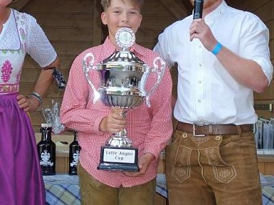 A boy holds a shiny trophy labeled 'Little Angler Cup' between two adults in traditional attire, at an event. The setting has a wooden backdrop and bottles.