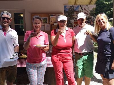 Five people in casual, sporty attire hold drinks and plates, standing outside near a table with a checkered cloth and posters on a wall behind them.