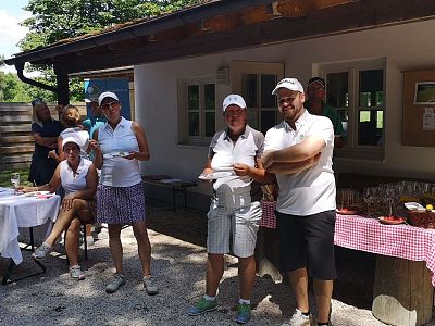 Four people in golf attire stand at a clubhouse smiling with drinks on a table nearby, set in an outdoor golf event scene.