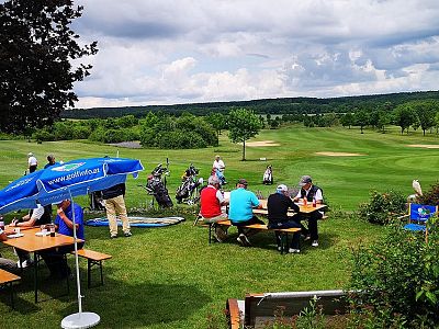 People sitting at picnic tables under umbrellas near a golf course, with golfers playing in the background under a partly cloudy sky.