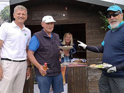 Three men and a woman at an outdoor event, with a food stall background, smiling and holding food items.