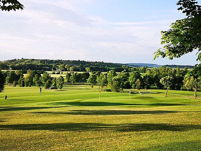 A green golf course with surrounding trees and distant hills, under a clear sky, creating a peaceful outdoor scene.