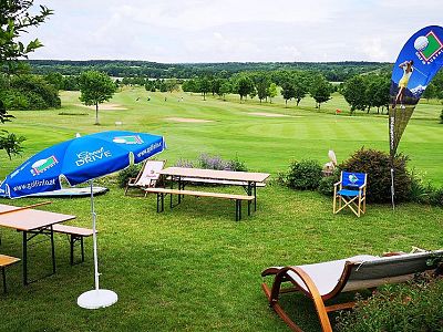 A golf course with green grass, trees, and a picnic area with tables, chairs, and a parasol. A flag and a deck chair are visible under a cloudy sky.