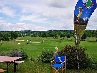 A scenic golf course with lush greenery, golfers playing in the distance, and promotional flags under a partly cloudy sky. Benches and seating in foreground.